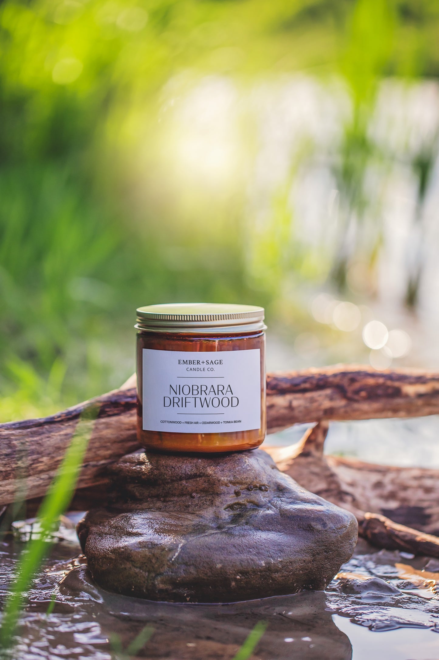 Niobrara Driftwood hand-poured Nebraska candle in amber glass jar on river bank on river rock with drift wood in the background and the sun shining through the grasses. Cottonwood, Fresh Air, Cedarwood, Tonka Bean scent