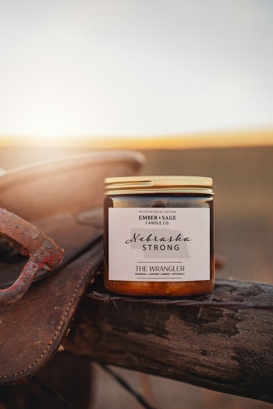Jar of 'Nebraska Strong' Wildfire Relief Edition candle in 'The Wrangler' scent on a wooden fence post with a leather saddle and branding iron in the background.