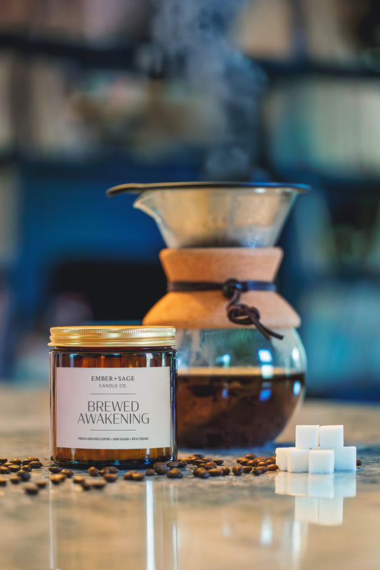 Brewed Awakening hand-poured Nebraska candle in amber glass jar with coffee beans, sugar cubes, pour-over coffee maker. Fresh ground coffee, raw sugar, rich cream scent.