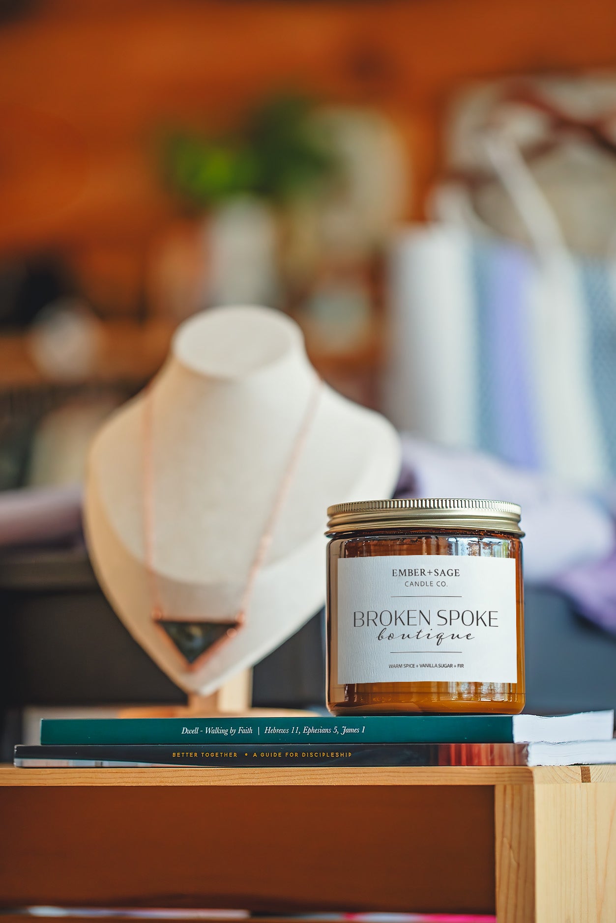Broken Spoke Boutique hand-poured Nebraska candle in amber glass jar in retail setting with a stack of devotional books, necklace and wooden wall in background. Warm Spice, Vanilla Sugar, Fir scent.