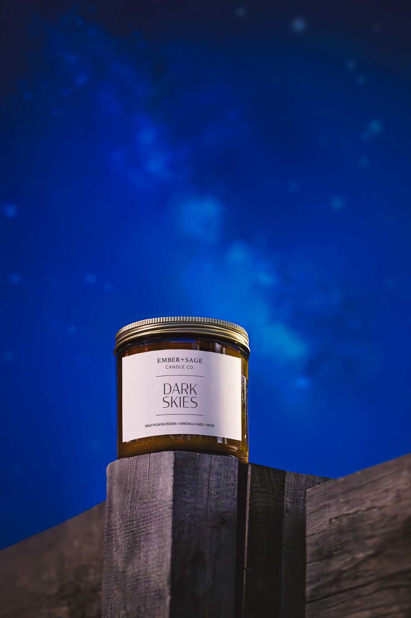Dark Skies hand-poured candle in amber glass jar for Nebraska International Dark Sky park sitting on cracked wooden post with night sky, stars, and Milky Way in the background. Winld Plum Blossom, Sandhills Sage, Musk scent.