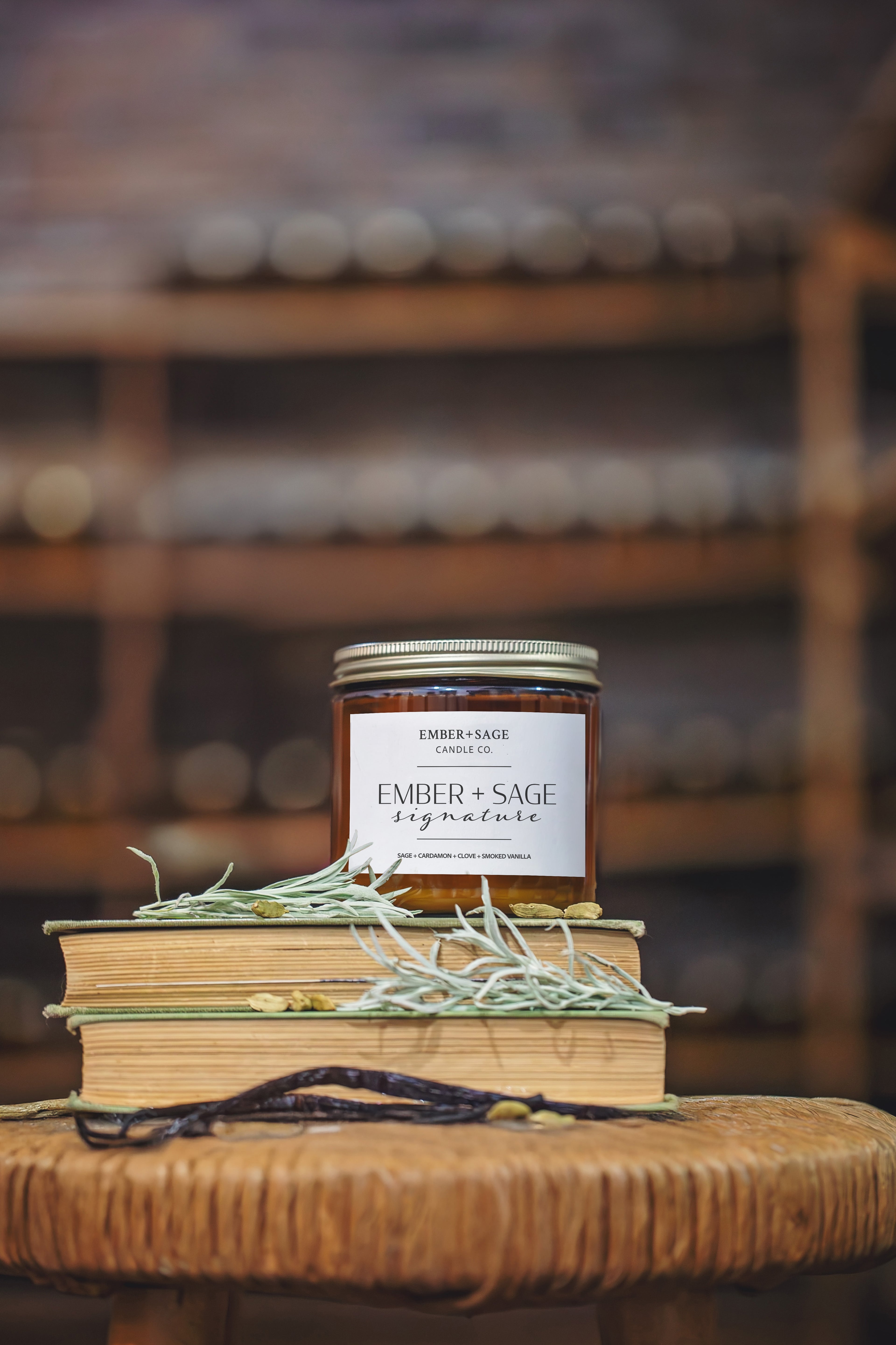 Ember + Sage Signature hand-poured Nebraska candle in amber glass jar sitting on books with sage, cardamon, and vanilla beans on a stool in a dimly lit brick vault with blurred our shelving. Sage, Cardamon, Clove, Smoked Vanilla scent. 