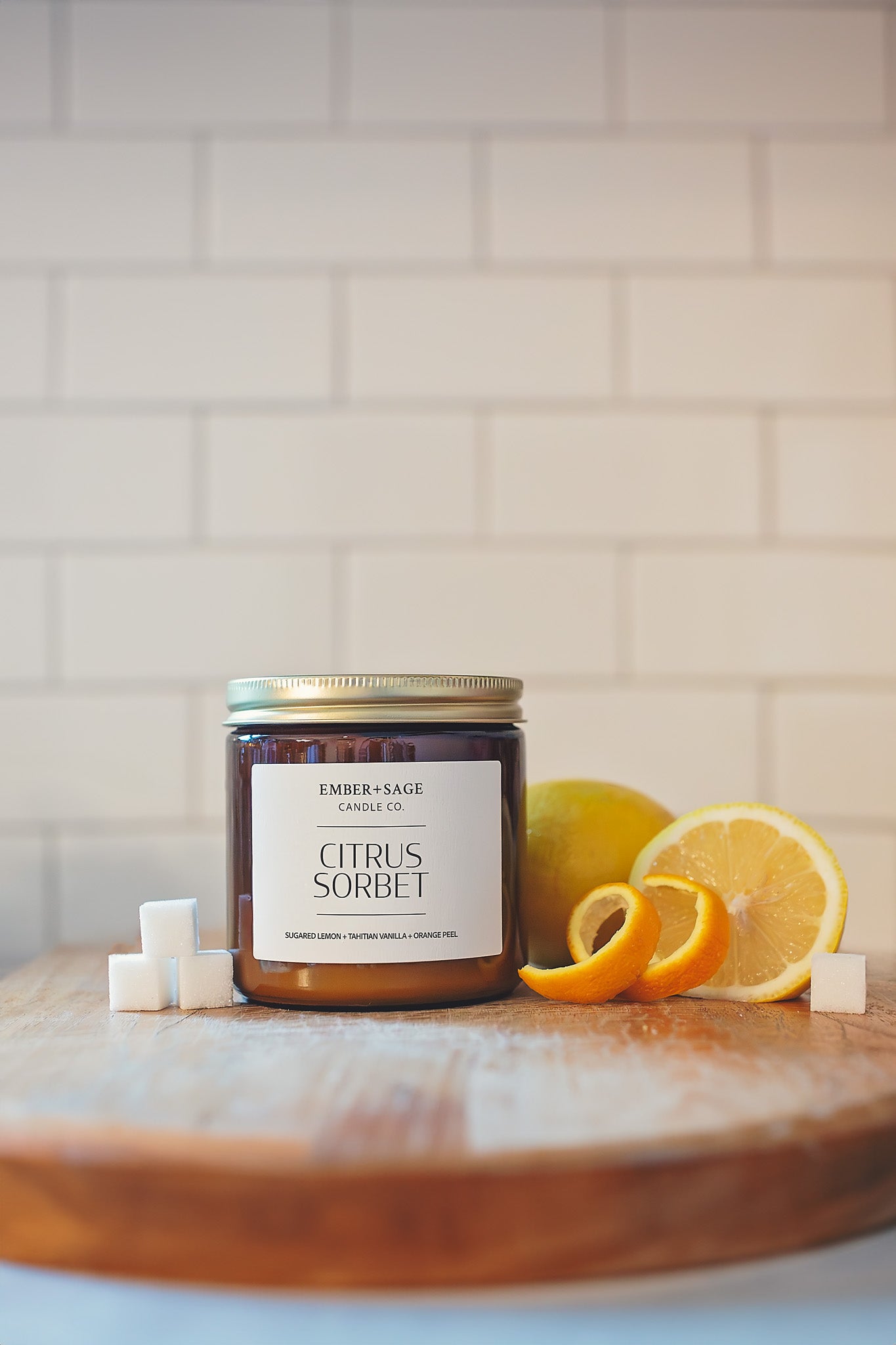 Citrus Sorbet soy/coconut Nebraska candle hand poured in amber glass jar with sugared lemon, Tahitian vanilla, orange peel scent on a wooden chopping block with lemon slices, sugar cubes, and spiral orange peel on clean white subway tile backdrop.