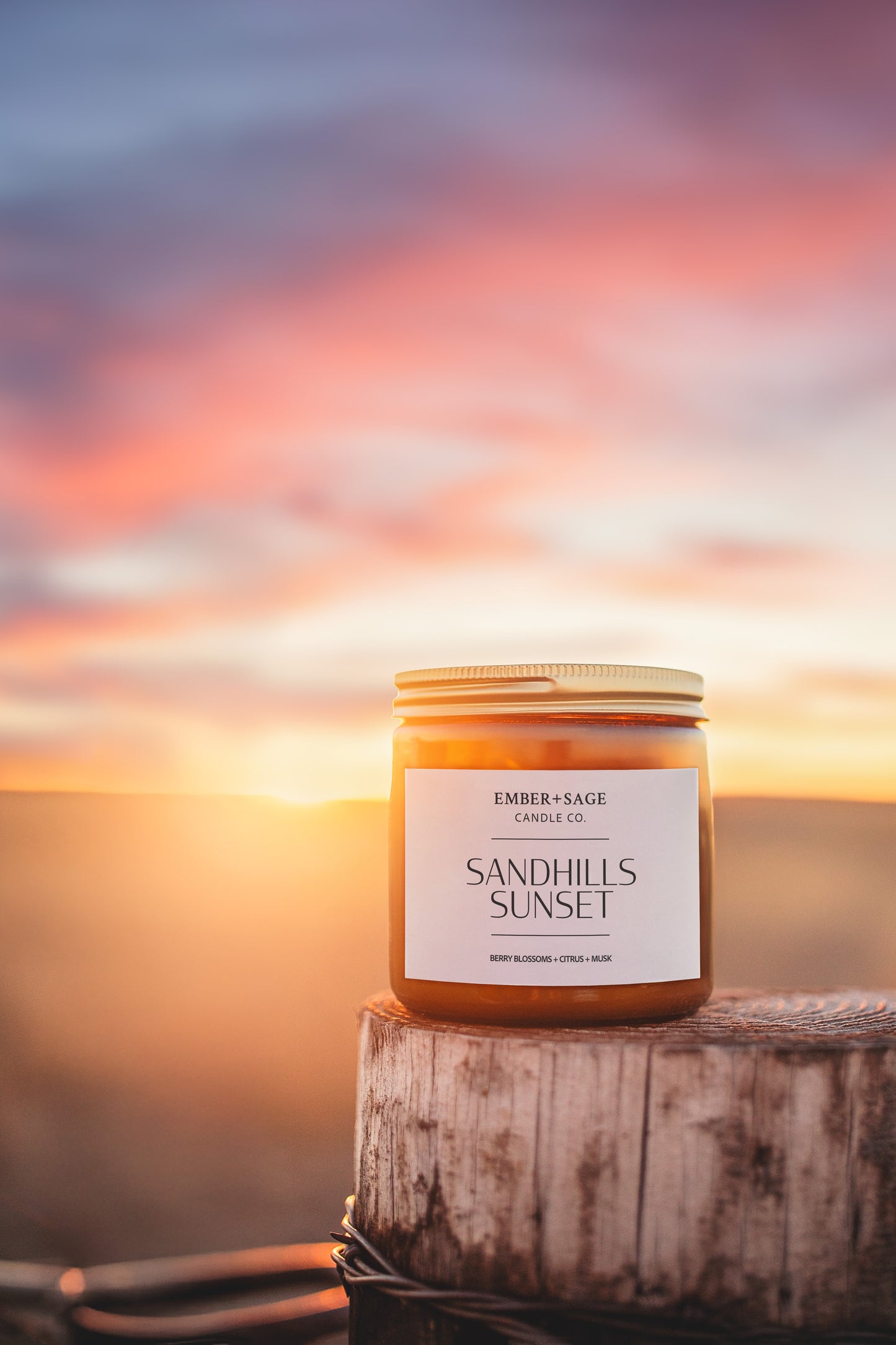 Sandhills Sunset hand-poured Nebraska candle on rustic fence post and barbed wire with beautiful sunset in distance. Berry blossoms, citrus, musk scent.