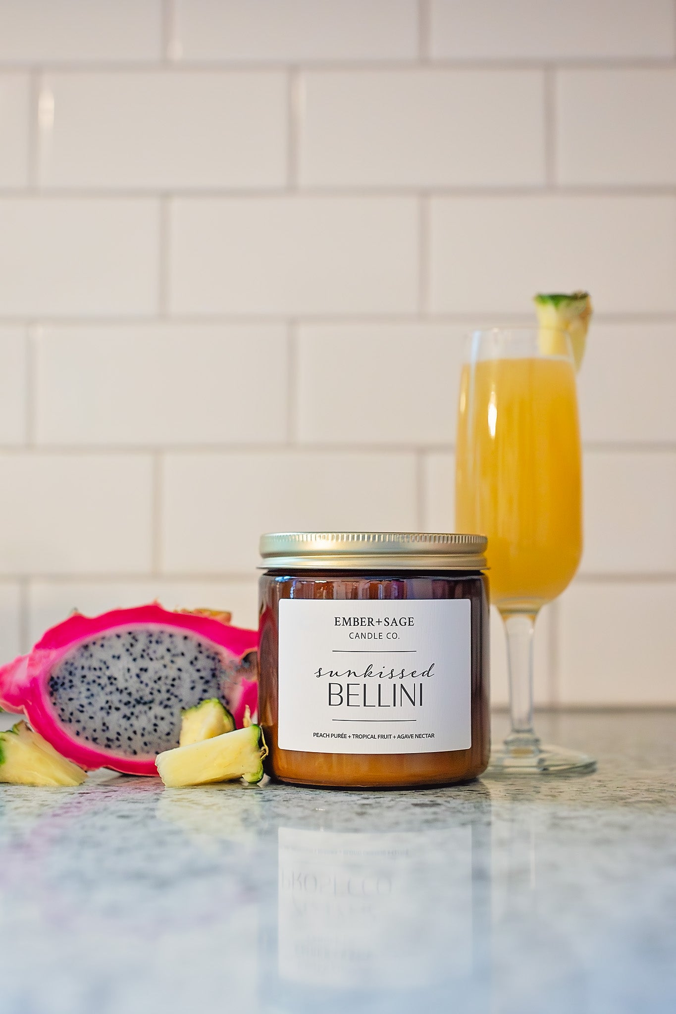 Sunkissed Bellini soy/coconut Nebraska candle hand-poured in amber glass jar with  peach puree, tropical fruit, agave nectar scent and peach Bellini drink, pineapple, and dragonfruit on shiny marble countertop and clean white subway tile backdrop.