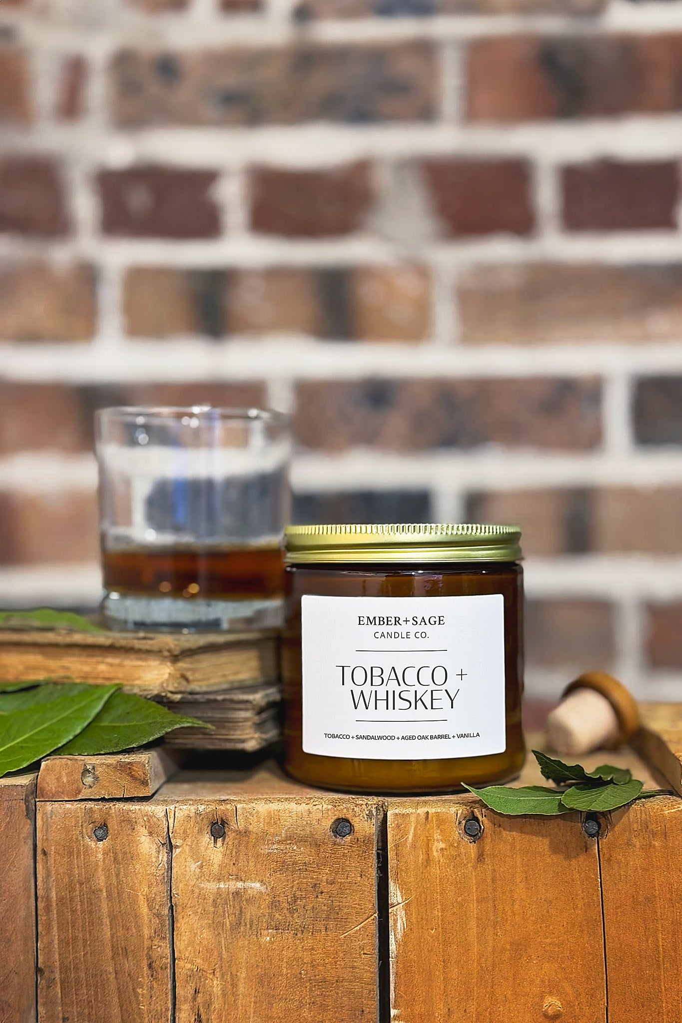 Tobacco + Whiskey hand-poured Nebraska candle in amber glass jar on wooden crate, tobacco leaves, cork and whiskey glass with rustic brick backdrop. Tobacco, sandalwood, aged oak barrel, vanilla scent.