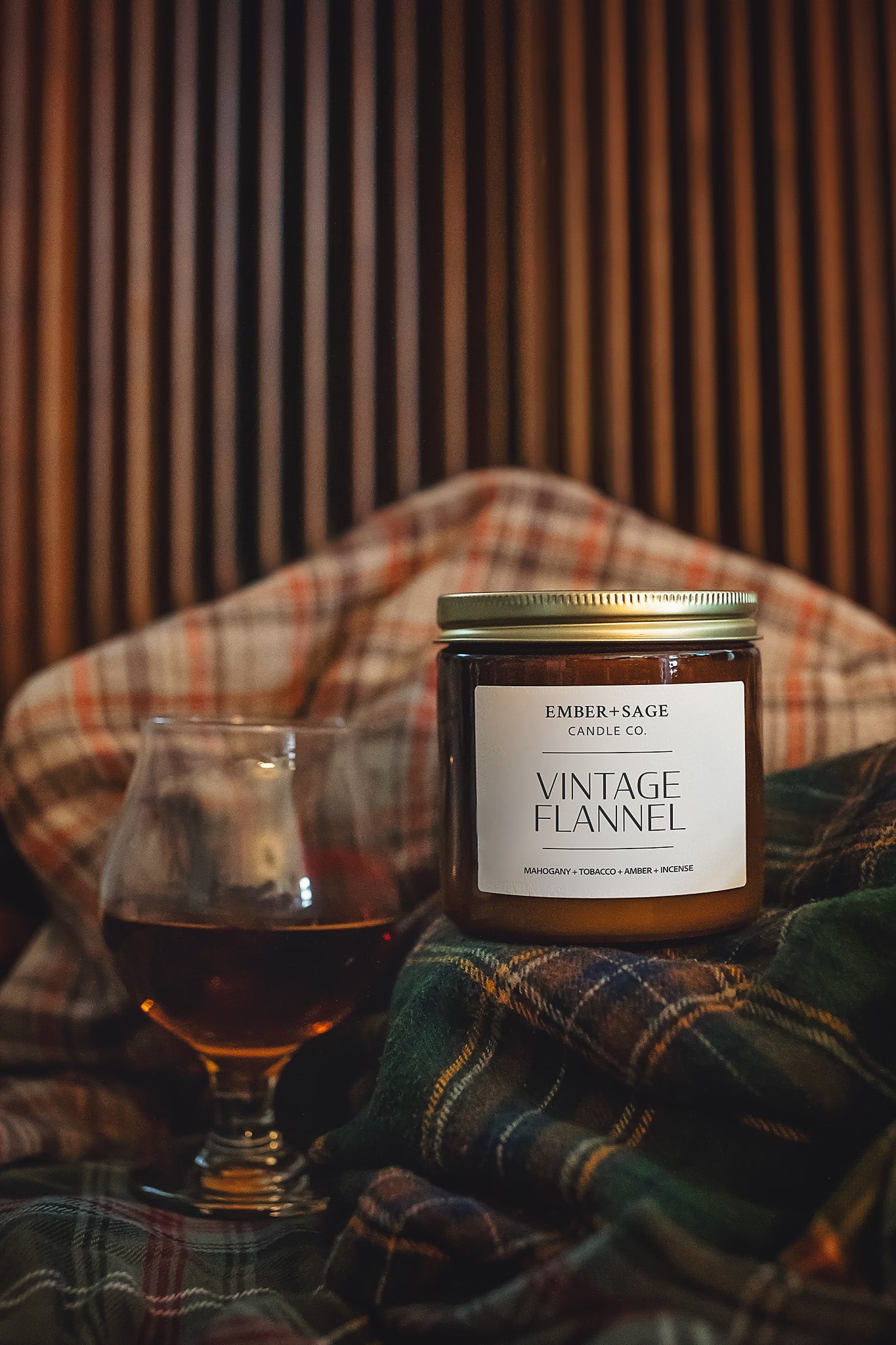Vintage Flannel hand-poured soy/coconut clean burning Nebraska candle with mahogany, tobacco, amber, incense scent on flannel shirts with glass of cognac and a wooden mid-century backdrop